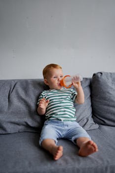 Child in stripped t shirt and denim pants sitting on sofa and drinking water from bottle
