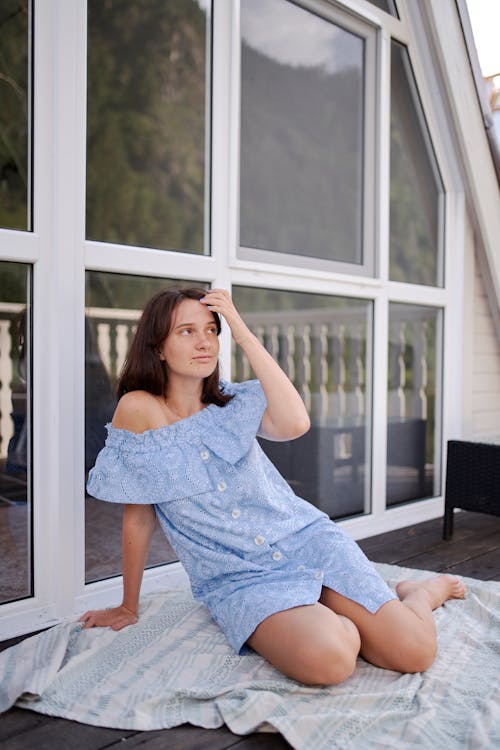 Full length young slim female in stylish sundress sitting on terrace floor and touching hair gently while looking away dreamily