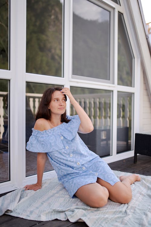 Free Full length young slim female in stylish sundress sitting on terrace floor and touching hair gently while looking away dreamily Stock Photo