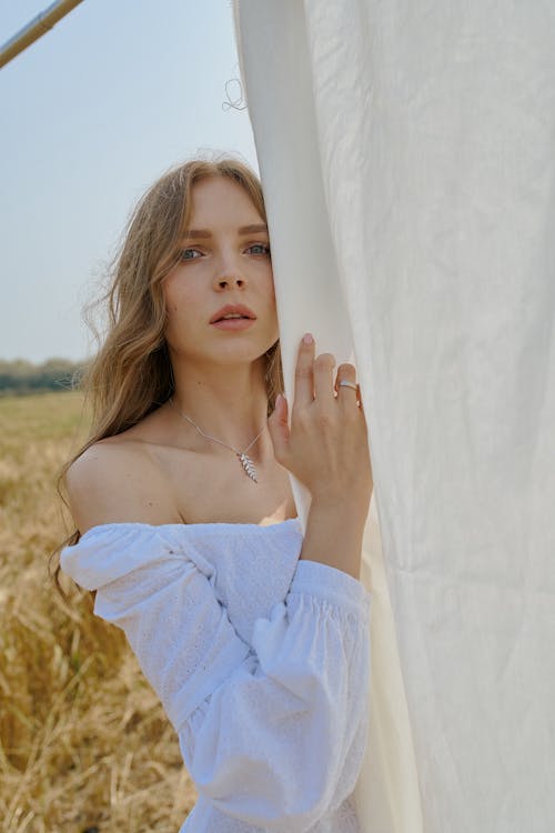 Serene young female wearing white dress with bare shoulders standing near hanging white sheet on sunny summer field and looking at camera