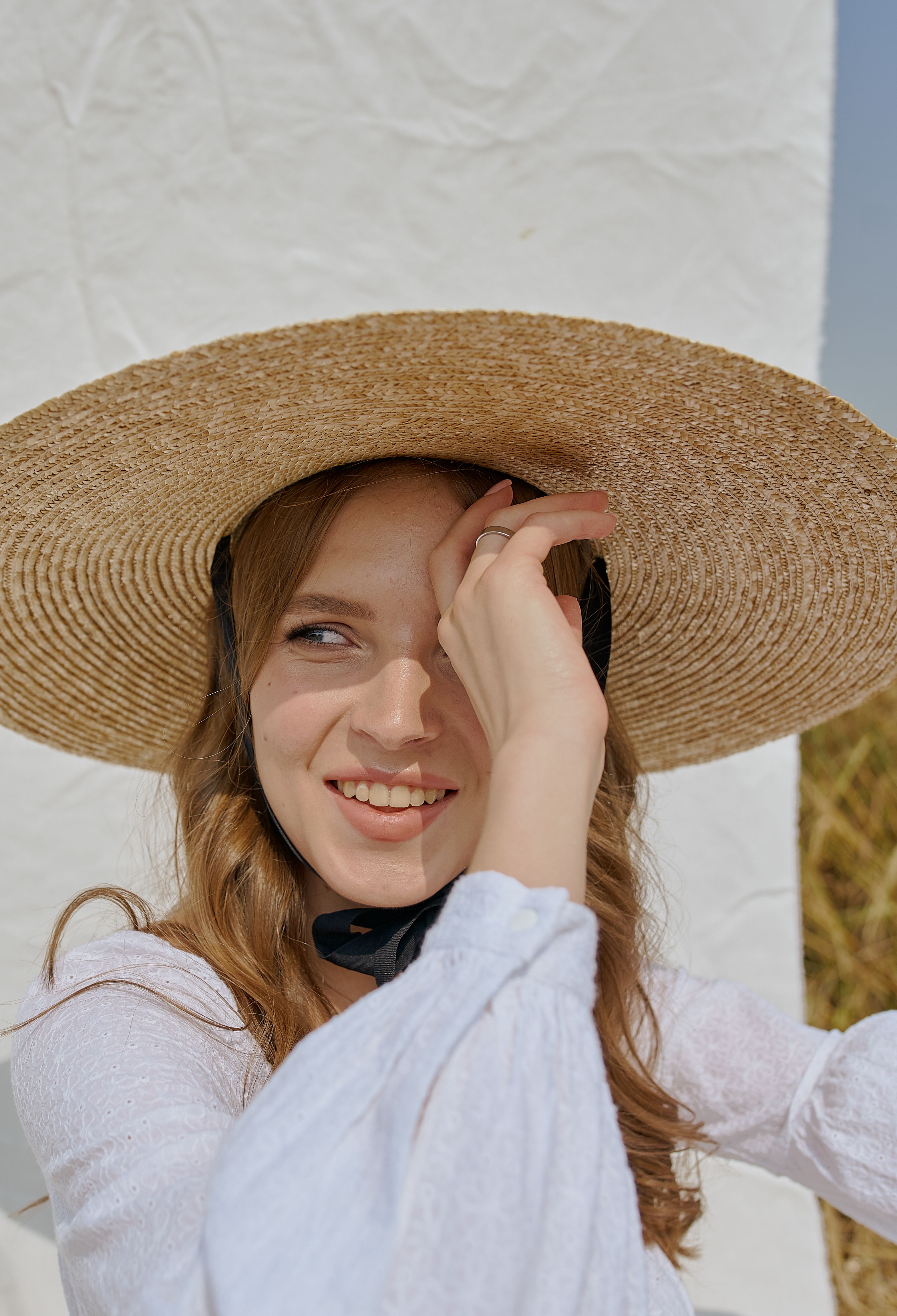 Smiling young female in straw hat standing against white sheet on sunny field while touching brim and looking away happily