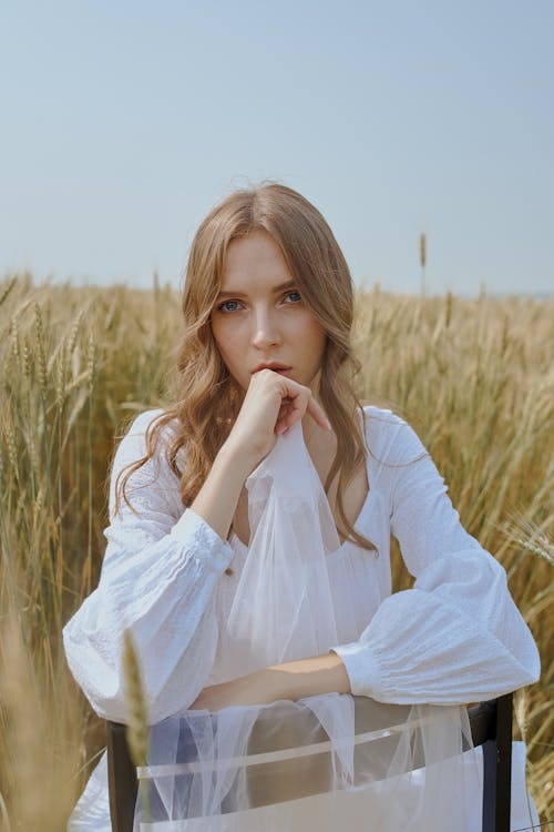 Pensive female with long wavy hair in stylish outfit touching chin and sitting on chair in field of wheat located in countryside in sunny day