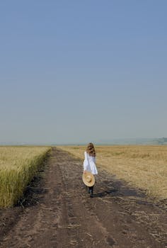 一名穿着休闲装的妇女沿着两块田地间的乡村小路行走，头顶是晴朗的蓝天