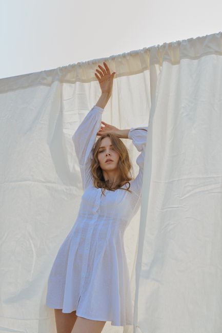 Charming model with long hair in trendy white dress standing with raised hand and touching head against white sheet hanging on street in daytime