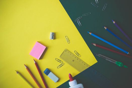 Top view of assorted school supplies like pencils, glue, and paper clips on a yellow and green background.