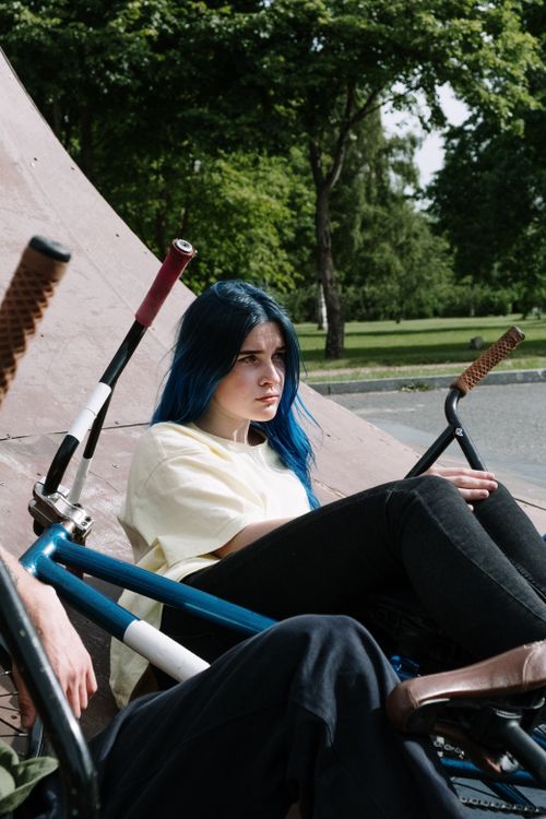 Free A woman with blue hair rests against a ramp with her bicycle at a skate park under a sunny sky. Stock Photo
