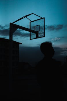 Silhouette of a person looking at a basketball hoop against a twilight sky.
