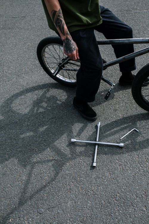 A cyclist crouched beside a bike on asphalt with hex keys and socket tools laid out on the ground, showing the tools and hands-on setup used for a home disc brake adjustment.