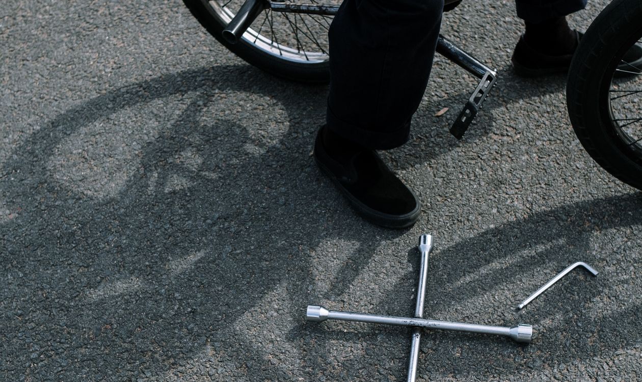 A cyclist crouched beside a bike on asphalt with hex keys and socket tools laid out on the ground, showing the tools and hands-on setup used for a home disc brake adjustment.