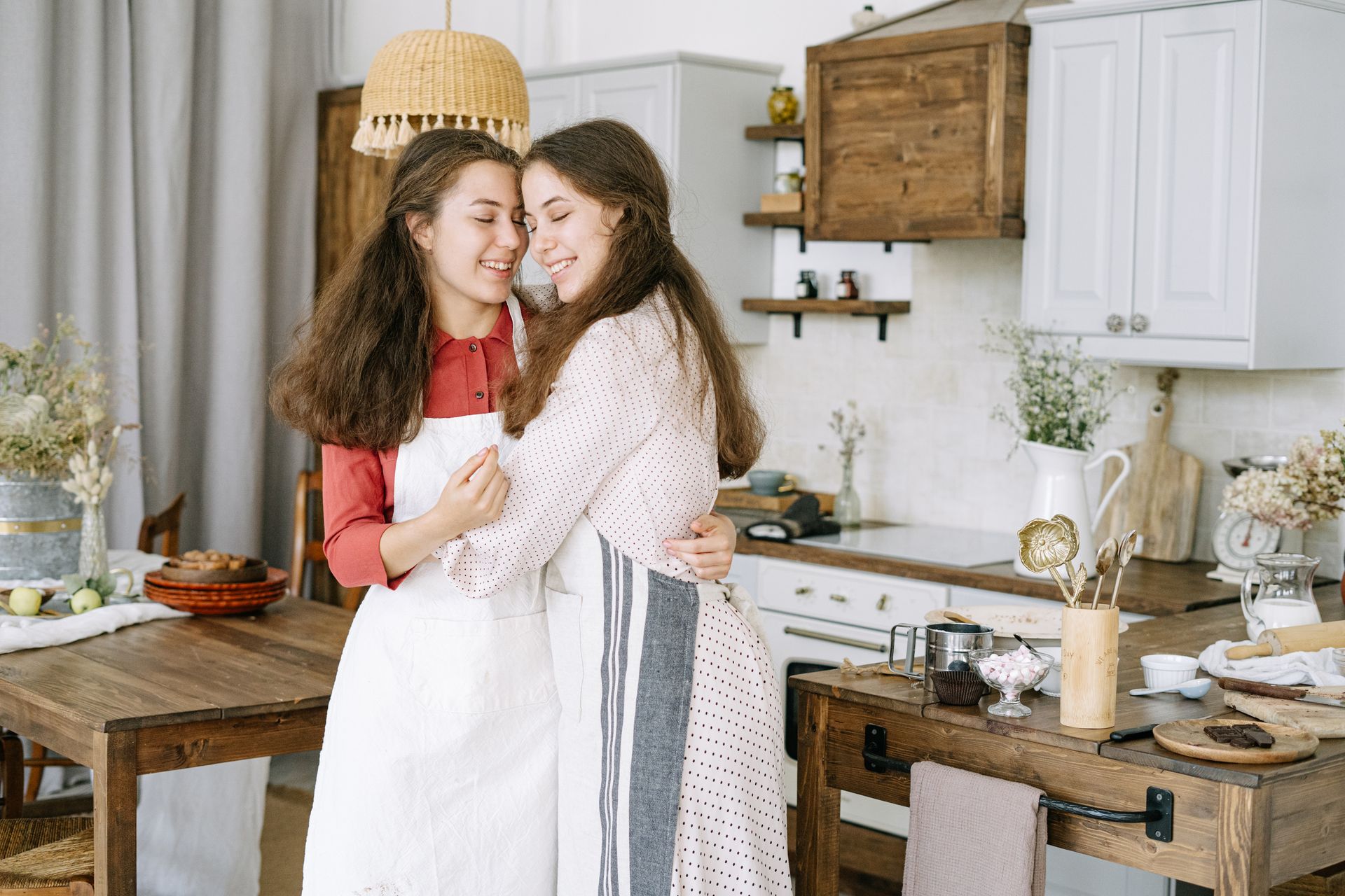 Two young women smiling and hugging in a rustic kitchen, captured indoors with a warm and welcoming atmosphere.