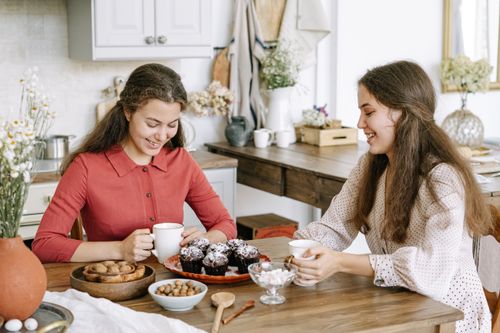 Free Two young women smiling and enjoying tea with muffins in a cozy kitchen setting. Stock Photo