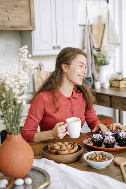 Free A smiling woman enjoying coffee and cupcakes in a cozy kitchen setting. Stock Photo