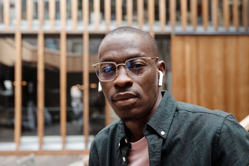 A Black man in his forties wearing clear-framed glasses and wireless earbuds in front of a wood-slatted building. Glasses are what a basic exam buys; the dilated exam is what protects his sight.