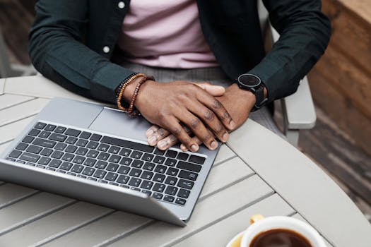 A person resting hands on a laptop, outdoors with coffee, suggesting a relaxed work environment.