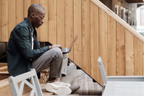 Free African American man sitting on stairs, working on laptop in a modern outdoor setting. Stock Photo