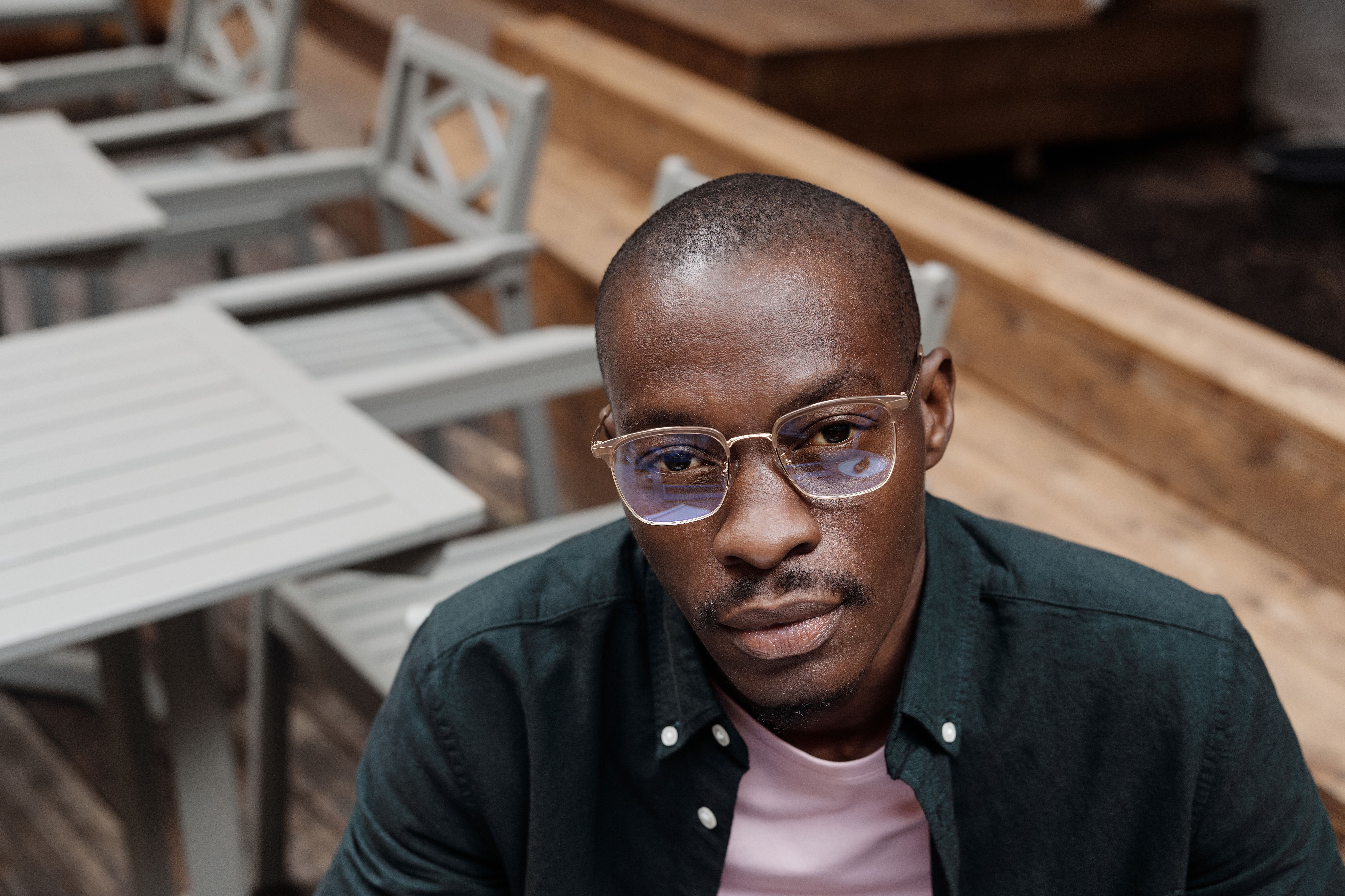 A fashionable man wearing eyeglasses poses outdoors at a cafe setting, exuding style and confidence.