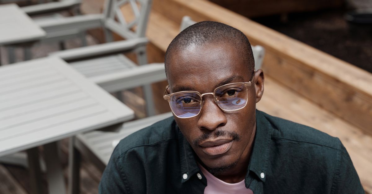 A fashionable man wearing eyeglasses poses outdoors at a cafe setting, exuding style and confidence.