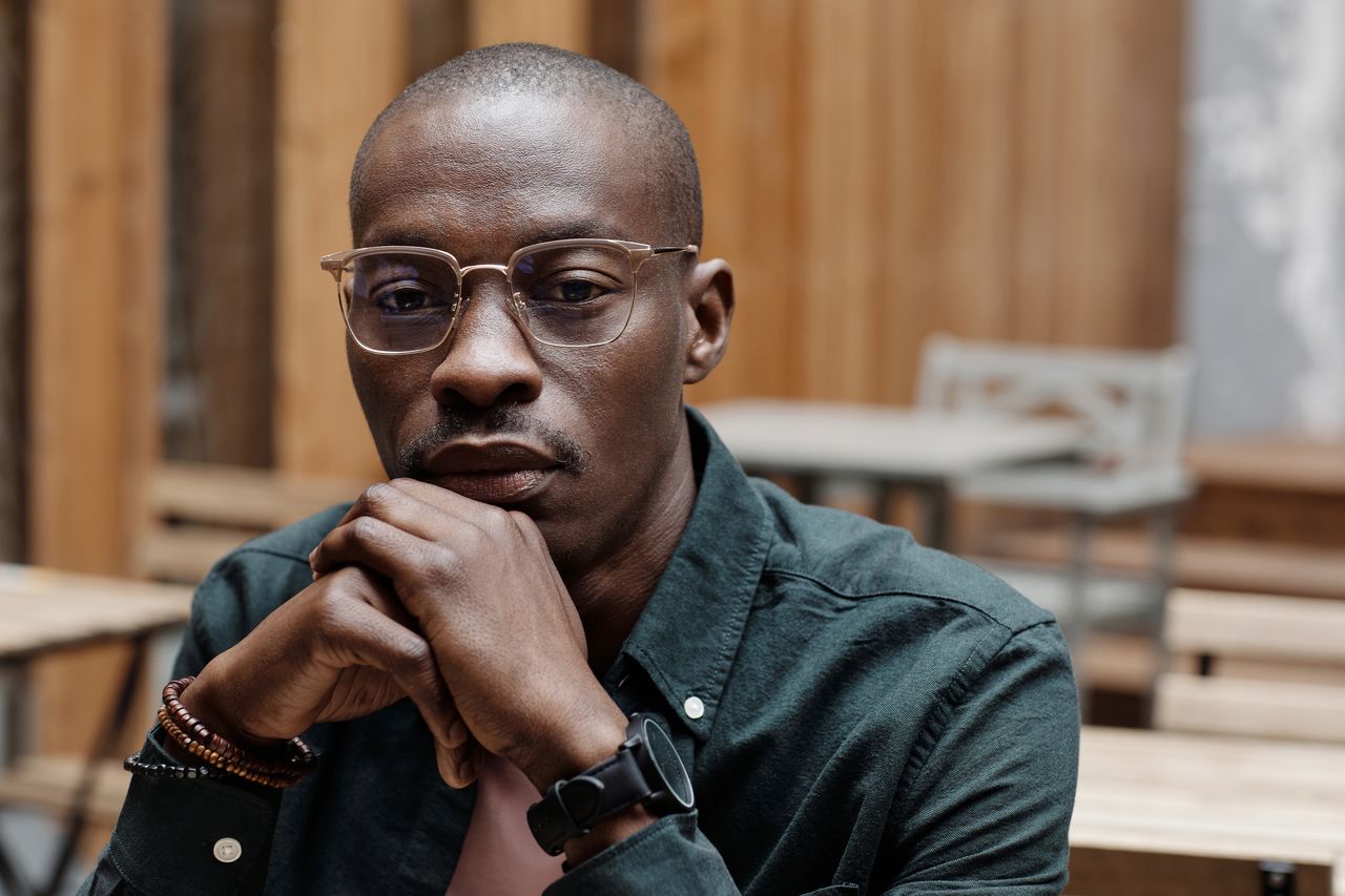 a contemplative man with eyeglasses sits indoors hands clasped