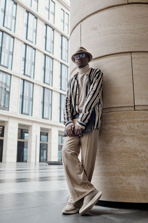 Free A fashionable man stands outdoors wearing a striped jacket, beige trousers, and a bucket hat, exuding trendy street style. Stock Photo