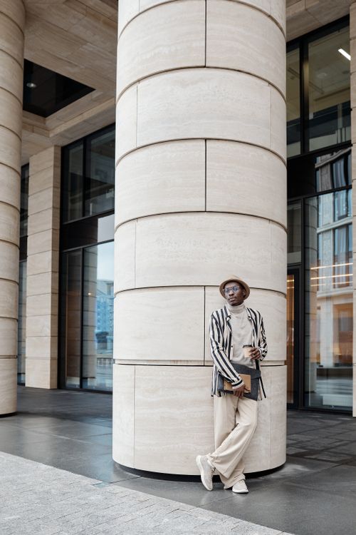 Free Fashionable man in striped attire poses by cylindrical column. Urban street style in modern architecture outside a building. Stock Photo