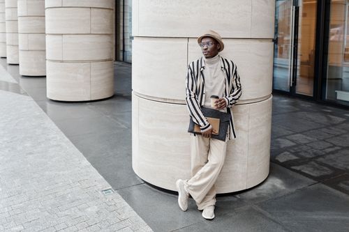 Free A stylish man in casual attire leans against large modern columns, holding a coffee and folder. Stock Photo