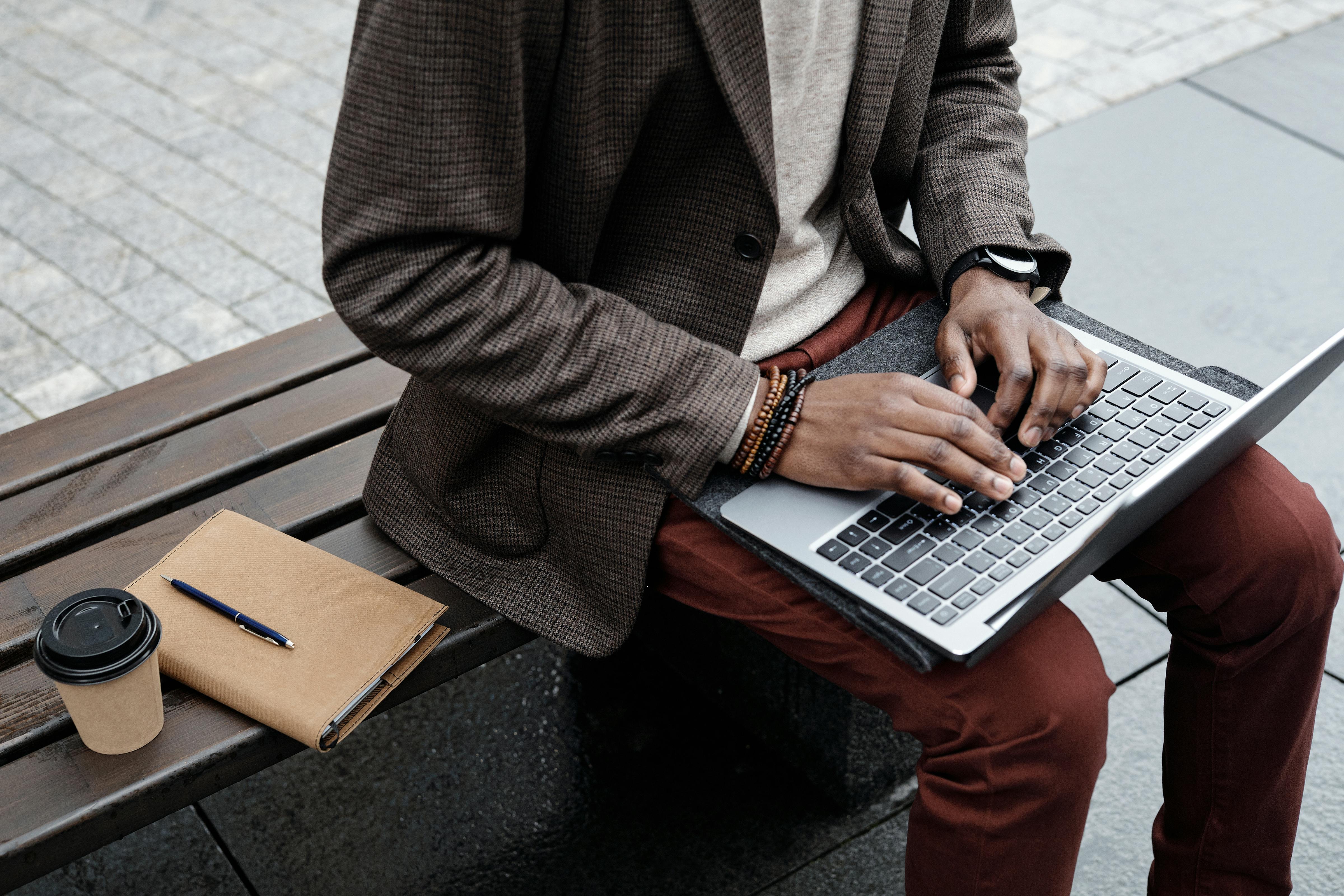 Man typing on laptop outside, with coffee, pen, and notebook on bench.