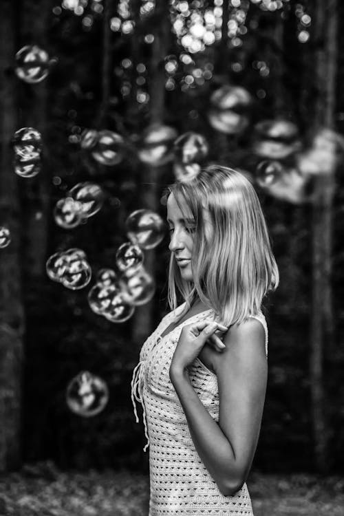 Black and white side view of peaceful female in summer dress surrounded by soap bubbles standing with closed eyes in woods