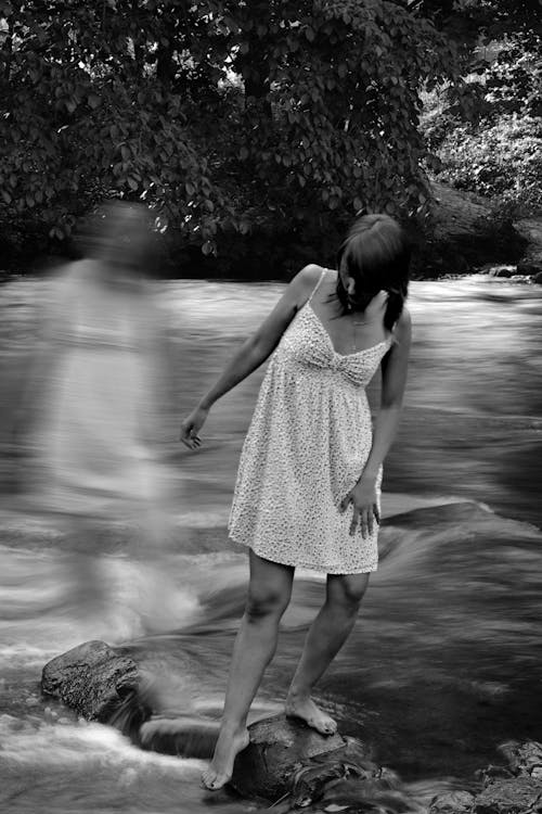 Free Black and white gentle female in dress standing on stone and touching water of river with leg Stock Photo