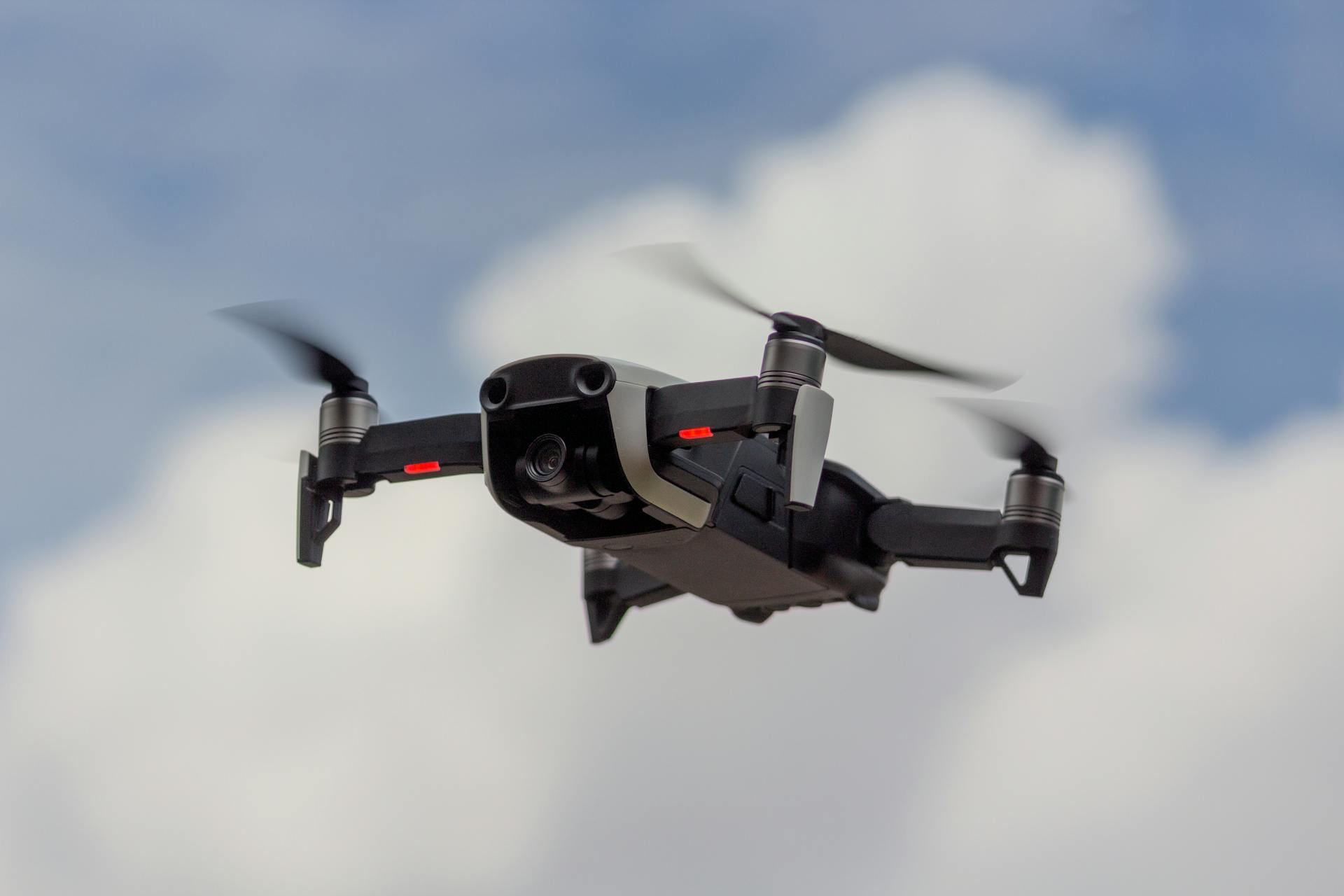 A sleek drone flying with rotating propellers against a blue sky with fluffy clouds.