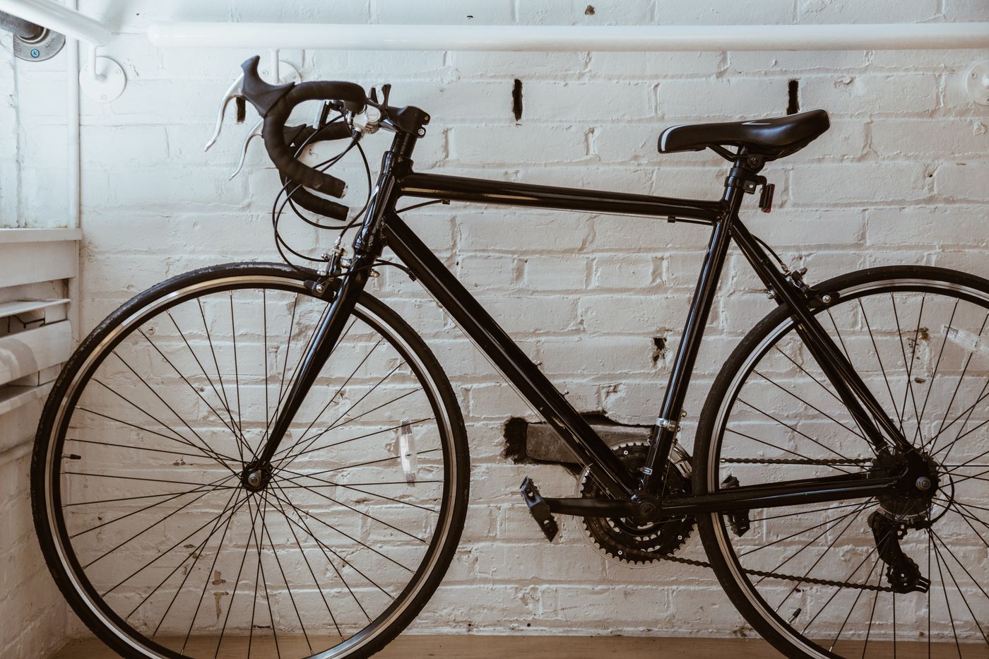A single black road bicycle parked/leaning against a rustic white brick wall in an urban outdoor setting. No people, no visible brand logos noted.