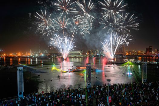 Spectacular fireworks light up the night sky over a bustling city, reflecting on the water below as crowds gather to watch.