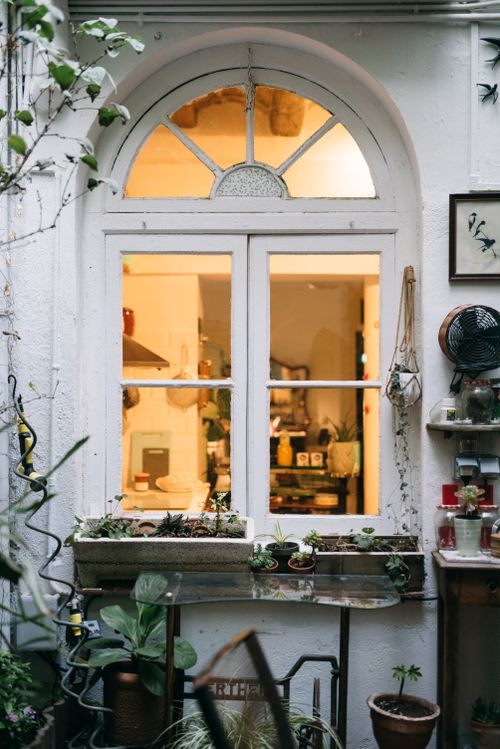 Free A charming garden view through an arched window with potted plants and a warm interior glow. Stock Photo