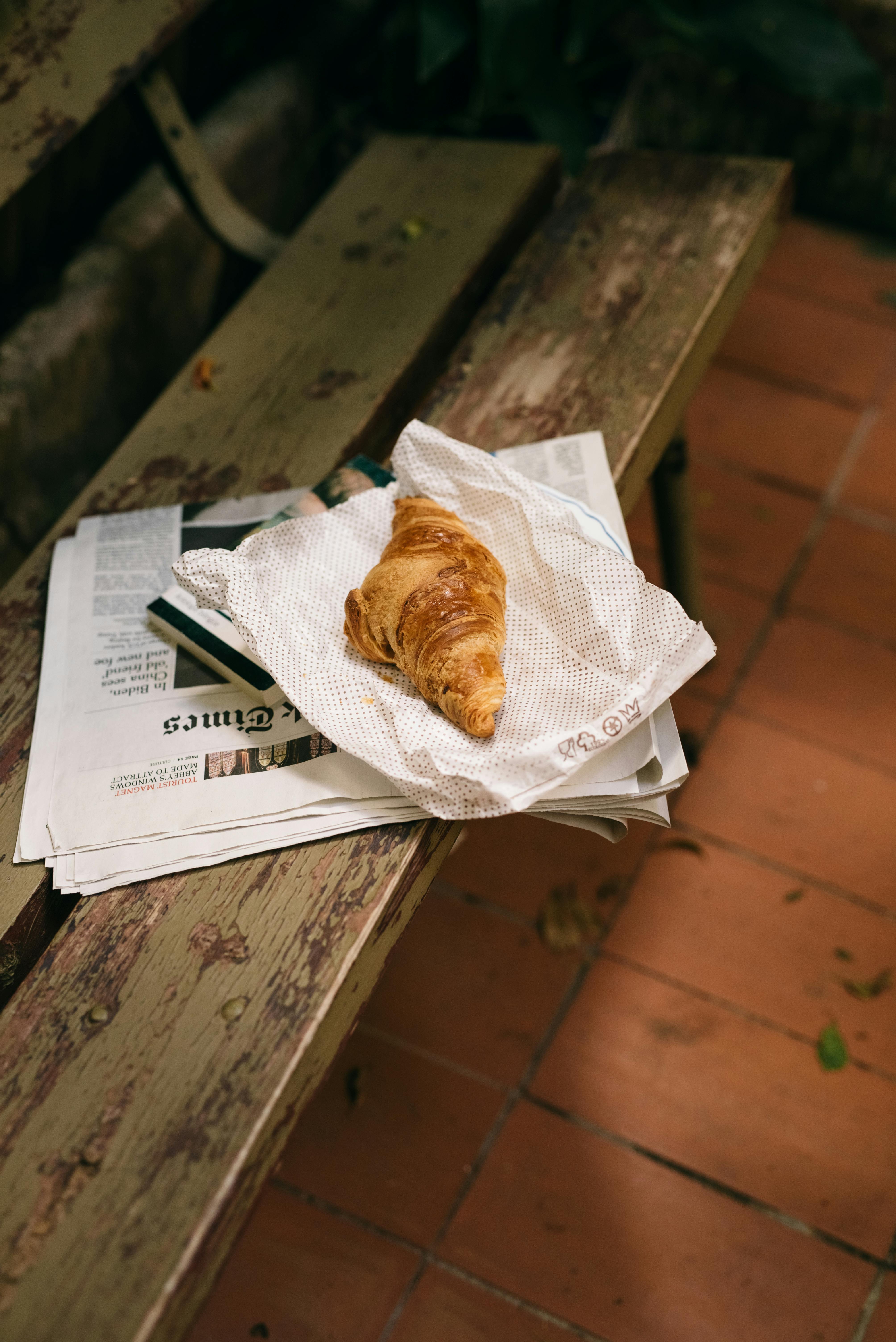 a croissant bread over a newspaper on a bench