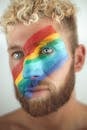 Crop handsome calm male with bright LGBT flag painted across face standing in light studio and looking away