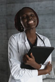 室内，一位身着白外套、面带微笑的非洲医生手持笔记本，肖像传达出专业与温暖