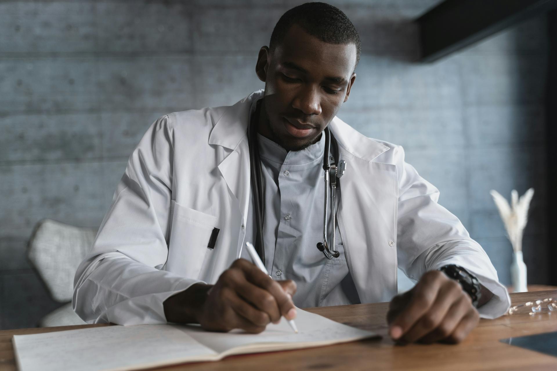 Black male doctor in a lab coat writing notes at a wooden desk in a medical office.