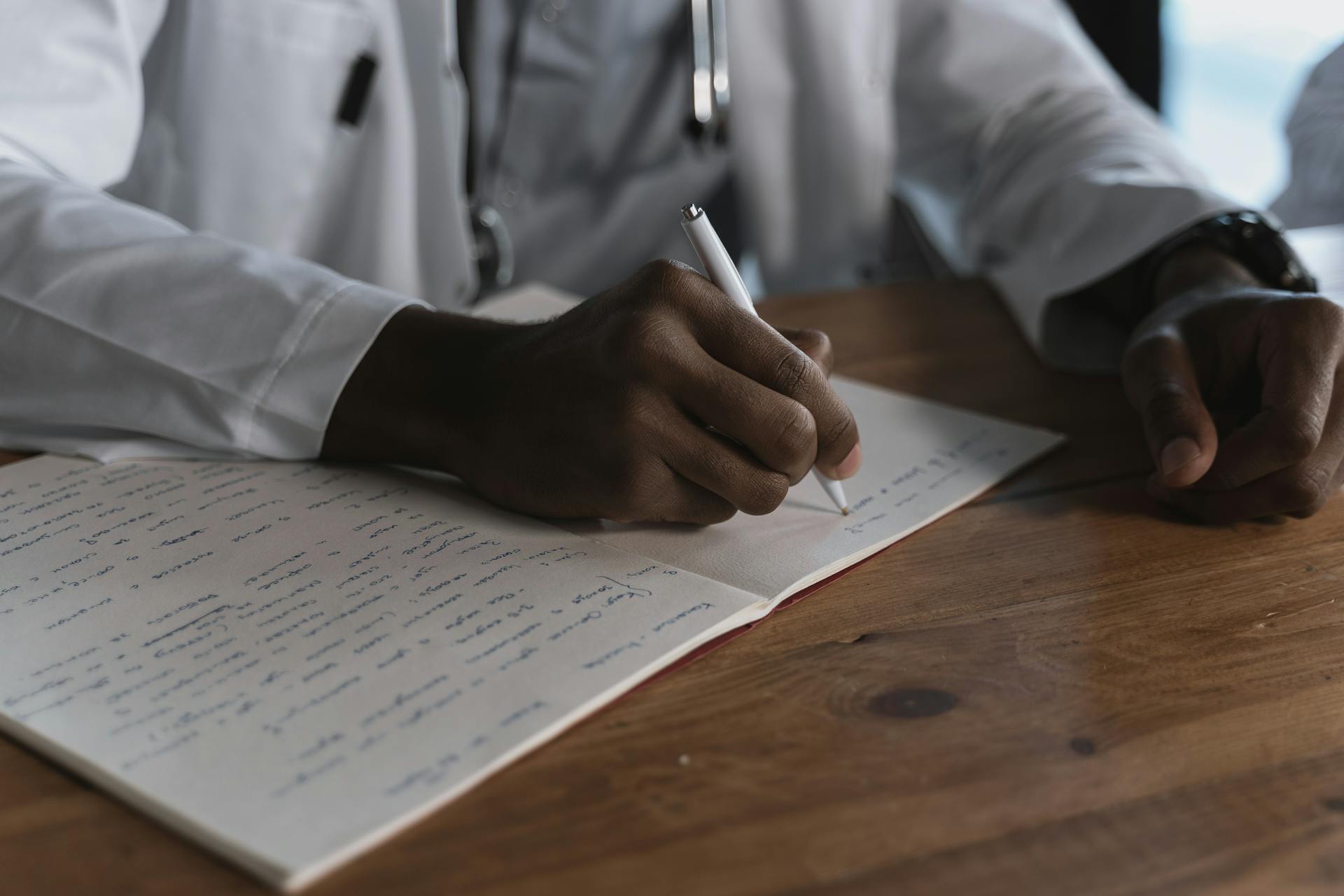 Close-up of a doctor writing notes with a pen in a medical notebook on a wooden desk.