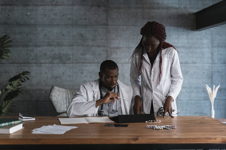 A Man And Woman With Stethoscopes  Wearing White Robes Looking At A Tablet On A Desk