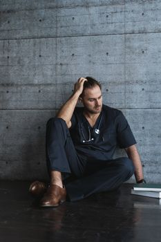 一位穿着手术服的医生坐在地板上，脖子上挂着听诊器，表情若有所思