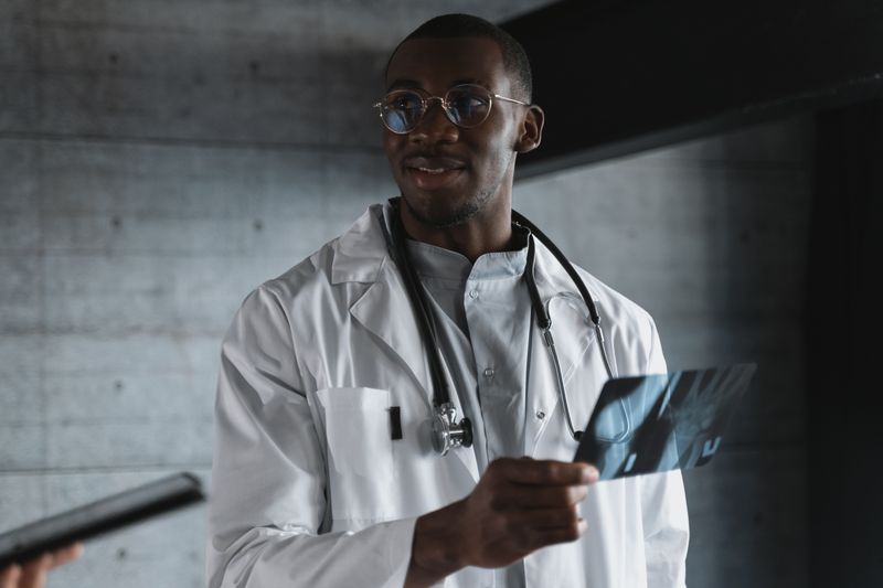 A Black physician in a white coat examines an imaging film, the kind of scan used to evaluate a persistent swollen lymph node and stage non-Hodgkin lymphoma.