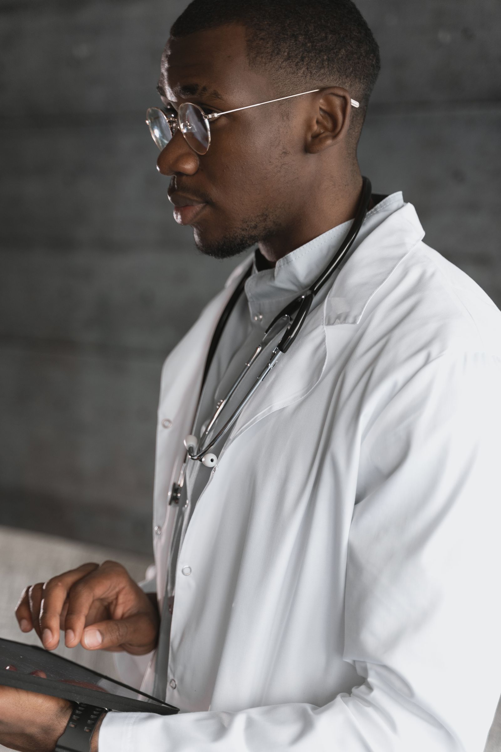 A Black physician in a white lab coat reviews records on a tablet in a clinical setting.