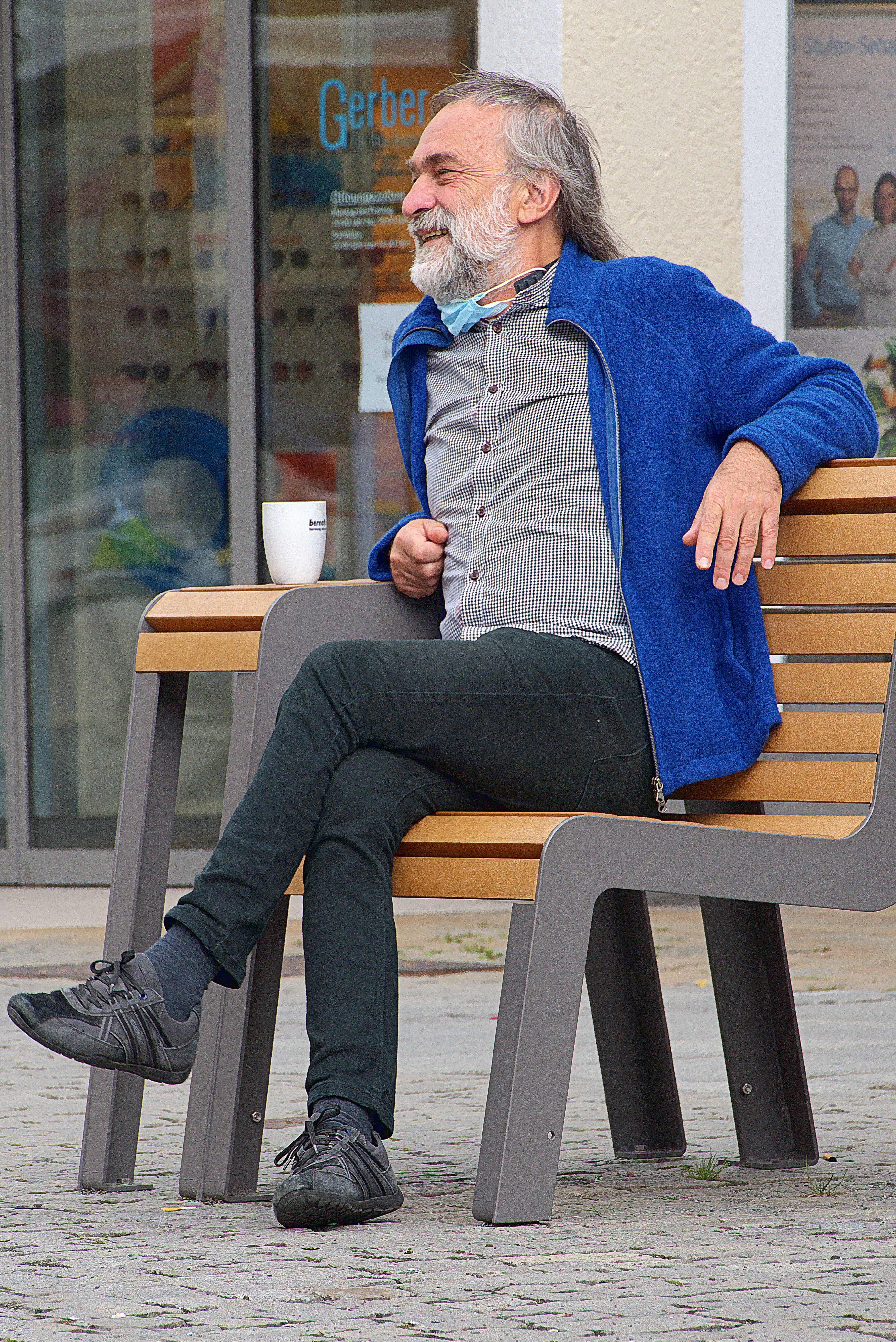 Smiling senior man sitting on a bench outdoors in casual attire with a coffee.
