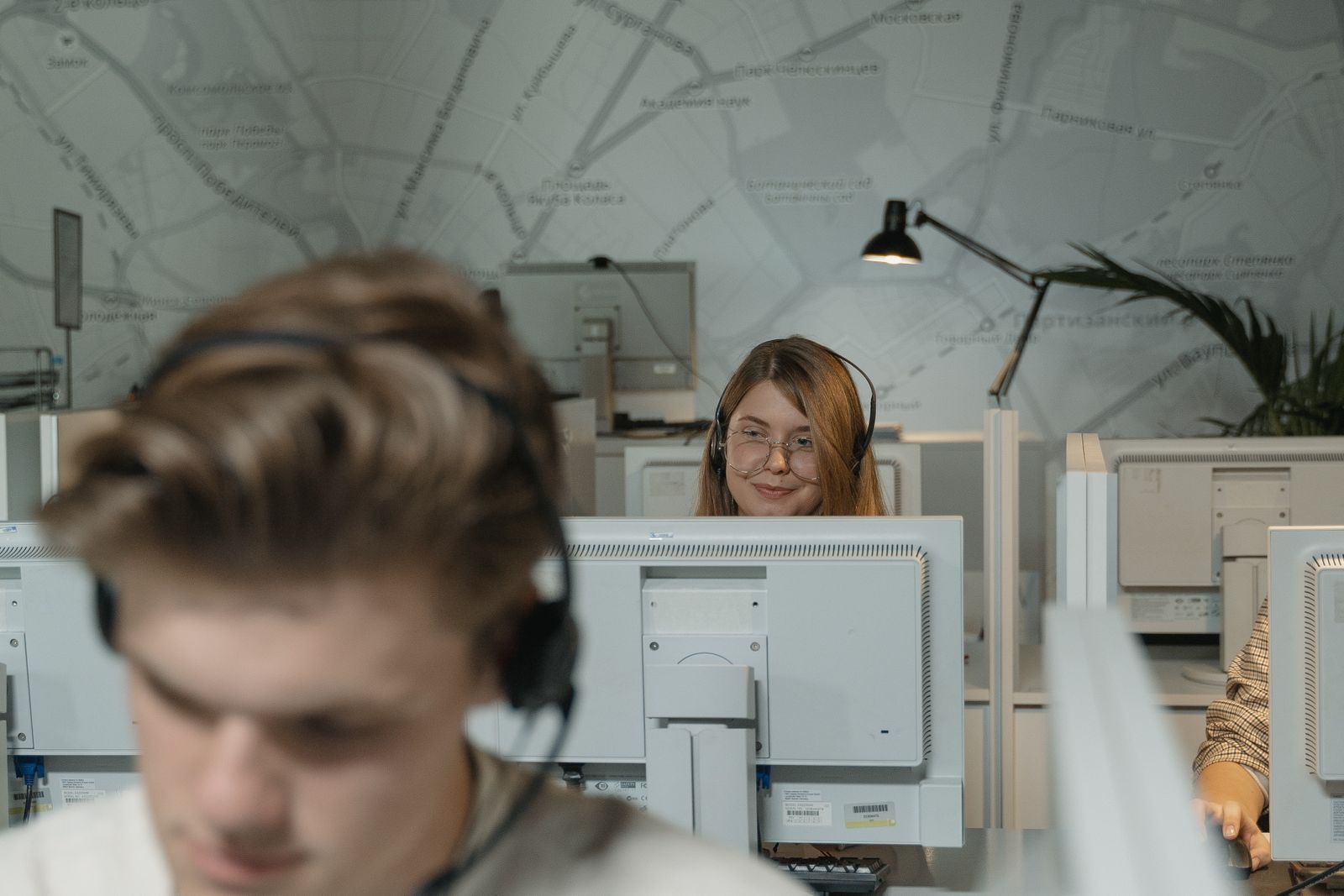 Due operatori di contact center davanti ai monitor sotto una grande mappa della città, in primo piano sfocata una persona con la cuffia.