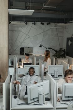 Modern office with diverse employees using computers and headsets in cubicles, providing technical support.
