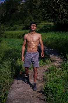 Muscular man shirtless walking outdoors on a sunny day in a natural setting.