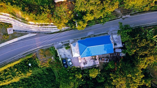 Aerial view of a rural house with a blue roof located in Moca, Dominican Republic, surrounded by lush green trees and a winding road.