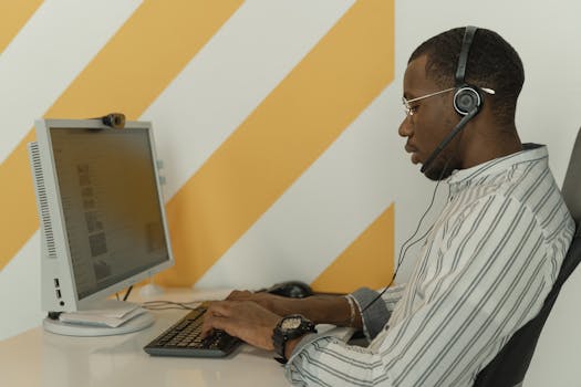 Side view of a black man wearing glasses and headset working at a computer in an office.