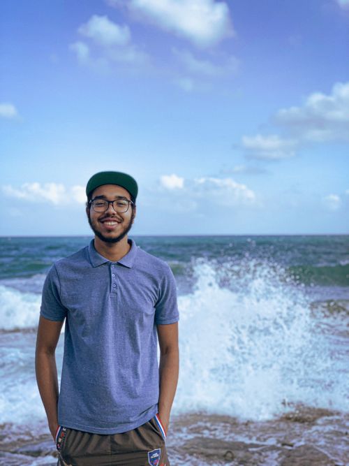 Free A young man in casual attire enjoys a sunny day by the ocean, smiling at the camera. Stock Photo