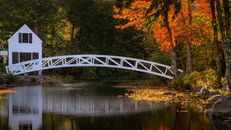 Scenic view of white arched footbridge over tranquil rippling river with rural cottage on coast in colorful autumn woodland