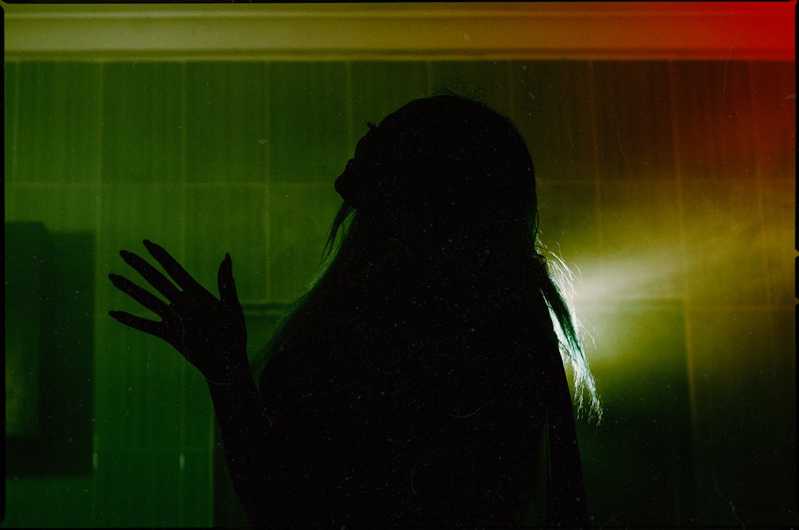 Side view silhouette of female throwing head back and raising hand with spread fingers while standing in dark room against dim green light